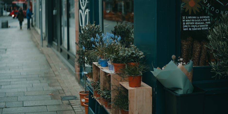 A display of various potted plants and floral arrangements outside a flower shop, with colorful flowers including blue, white, and green foliage arranged in terracotta and blue pots placed on wooden s