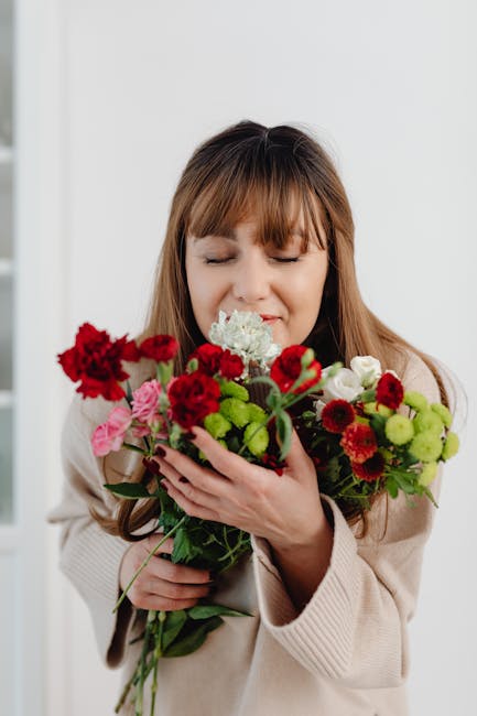 A woman with light brown hair and bangs, wearing a cream-colored cardigan, is holding a lush bouquet of fresh flowers close to her face with a gentle, contented expression and her eyes closed. The bou