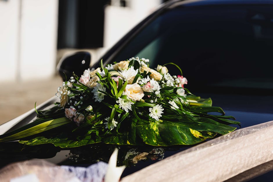 A floral arrangement of fresh white roses, daisies, and small pink accents, artfully arranged with green foliage and leaves, rests on the hood of a sleek black car. The bouquet is wrapped loosely with