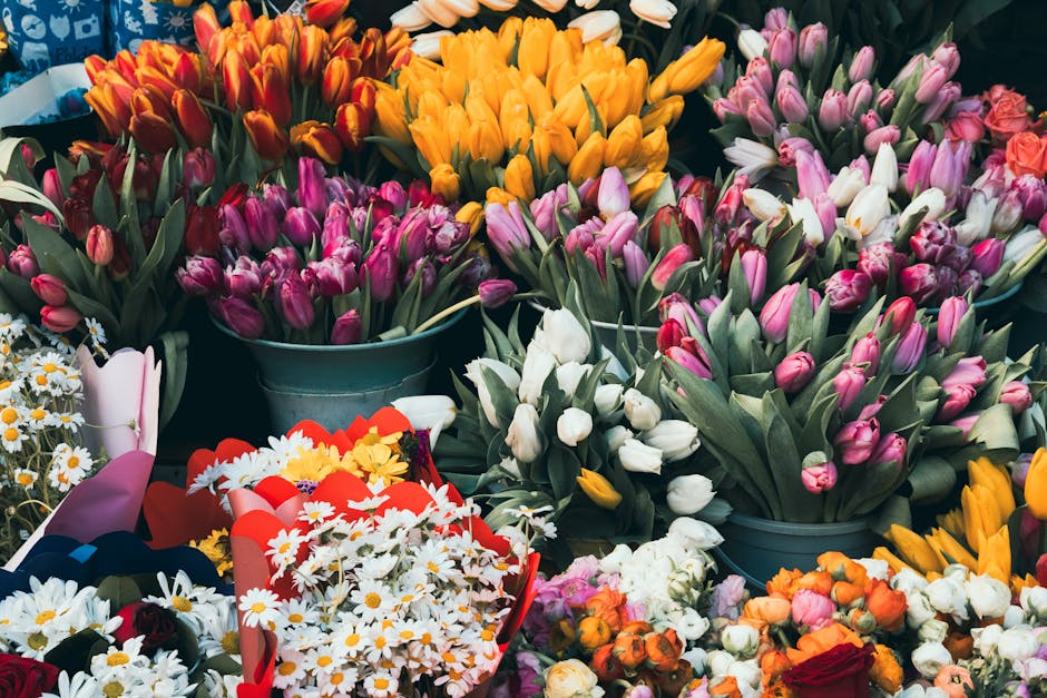 A vibrant display of various fresh flowers including pink, purple, white, yellow, and orange tulips arranged in multiple metal buckets, with some bouquets wrapped in decorative paper and ribbons. Dais