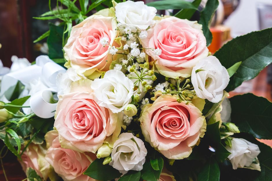 A bouquet of fresh flowers arranged with pale pink roses and white lisianthuses, accented by small white baby's breath and lush green foliage. The roses have gently spiraled petals with a soft, velvet