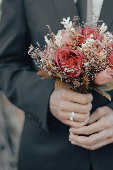 A close-up of a person holding a floral bouquet with both hands, featuring large, deep pink roses as the focal point, accented by smaller white and beige flowers, dried foliage, and sprigs of lavender