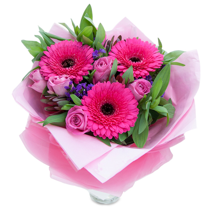 A carefully composed pink bouquet photographed on white, where three fuchsia Gerbera daisies dominate the view with their wide, perfectly formed petals and deep, near-black centres that lend dramatic contrast. Nestled around them are a handful of rosebuds in a muted lavender-pink or dusty-rose tone; their velvety, inward-curving petals read as fresh and slightly fragrant, adding a romantic softness to the arrangement. Sprigs of purple filler-statice or limonium-hover like tiny constellations between the larger flowers, contributing fine texture and a cool tonal counterpoint. The supporting greenery, a mix of broad, glossy leaves similar to salal or eucalyptus, gives the bouquet structure and a dewy, just-cut-from-the-garden quality. The wrapping is part of the composition: a pale pink matte tissue forms an understated base layer while an outer, semi-sheer fuchsia-pink cellophane with gentle crinkles catches the light and amplifies the pink theme; the bouquet is folded and tied with care so it reads as both elegant and approachable. At the base the clear glass vase's top rim is visible, underscoring that the arrangement is ready to go straight into the home. Bathed in soft, neutral light, the interplay of textures-silky rose petals, papery statice, crisp gerbera rays and glossy foliage-feels tactile and inviting. This bouquet is the kind of piece that would lift a bedside table, serve as an intimate centrepiece for a small celebration, or arrive as a warm, celebratory greeting for someone in or near Becontree Heath, Dagenham or Barking who deserves a bright, thoughtful gesture.